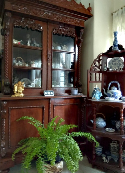 A Boston fern sits on the marble floor near two elaborately carved and polished teak display cabinets.